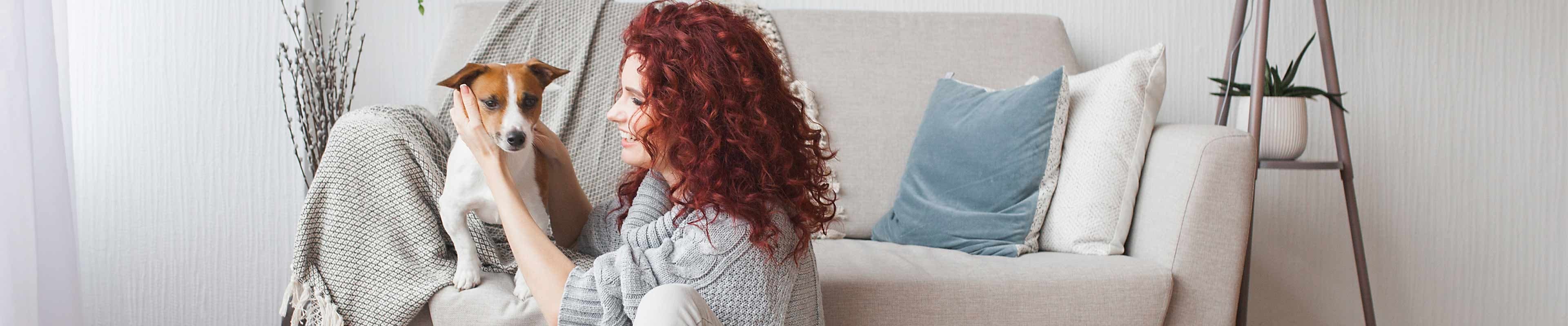 A red-haired white woman cuddles a small terrier breed dog that is sitting on a beige couch in a Scandinavian-style minimalist apartment.