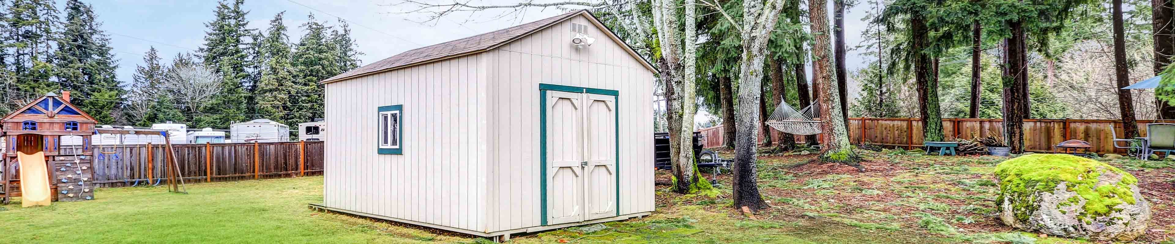 A white backyard shed with vertical siding and teal blue painted trim around the doors and on the single window on the side built in a mowed, fenced-in backyard with a kids' slide and a hammock. There is a trailer park in the background behind the fence.