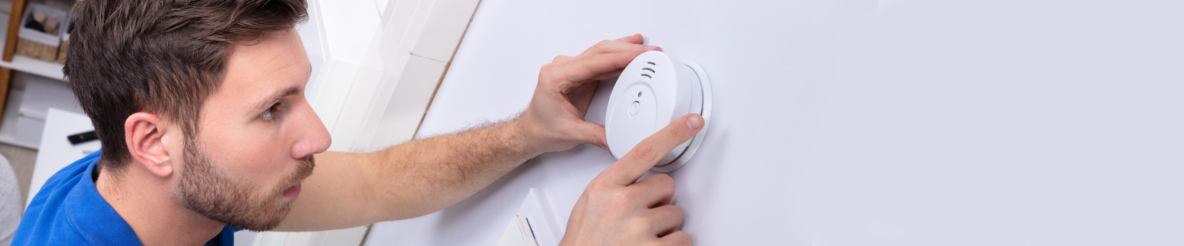 A white man with brown hair and closed-cropped beard wearing a blue shirt is installing a smoke detector on a white wall.