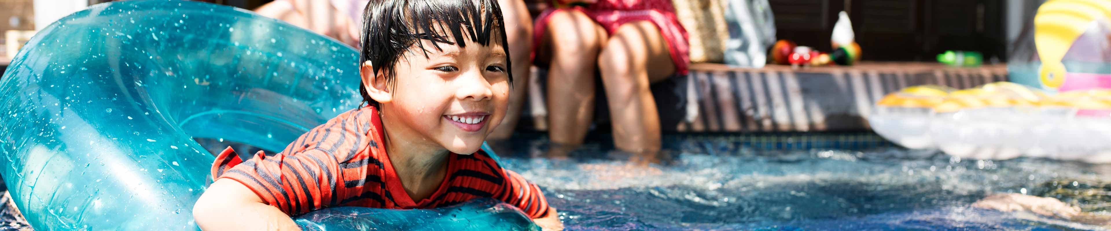 A smiling Southeast Asian boy in a red and black striped t-shirt swims in his family's pool using an inner tube to float while his mother and father sit on the edge of the pool watching him.
