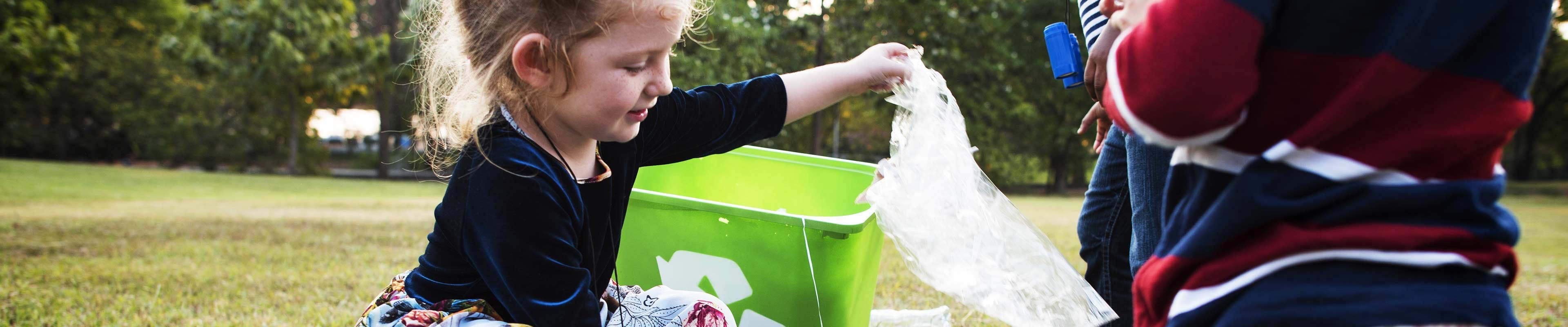 A little blonde white girl wearing a blue velvet dress sorts plastic trash on a lawn into a green recycling bin.