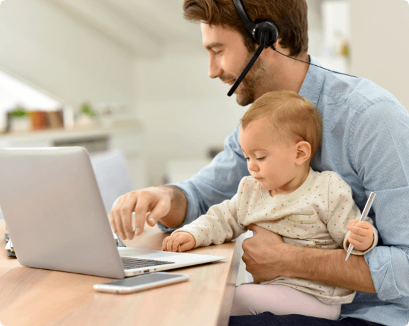 Father holding his infant at a table while working on a laptop.  