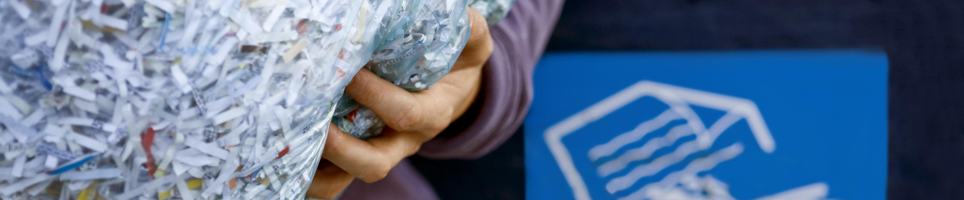 A person holding a bag of shredded documents in front of a shredded paper disposal bin.