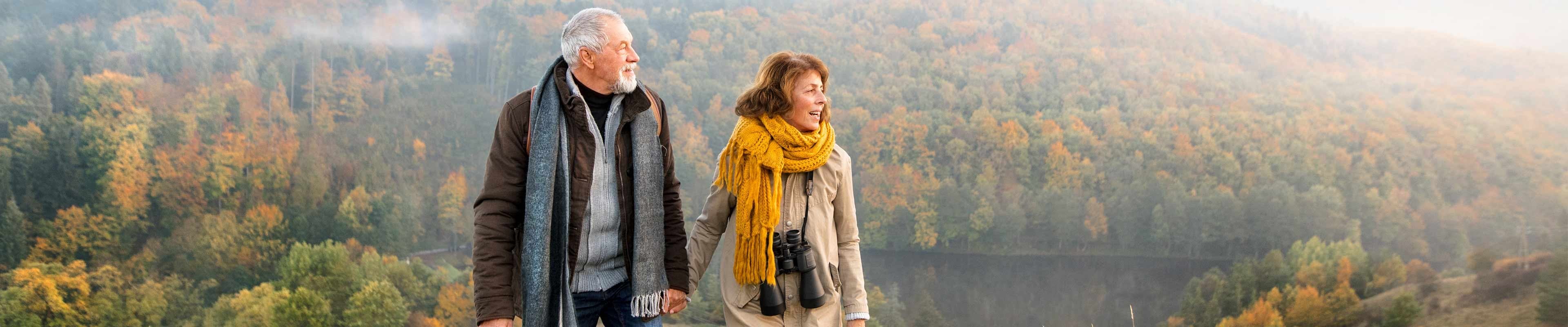 A elderly white man with a short-cropped white beard and an elderly white woman with dyed light brown hair holding hands on a mountain overlooking a lake in the autumn.