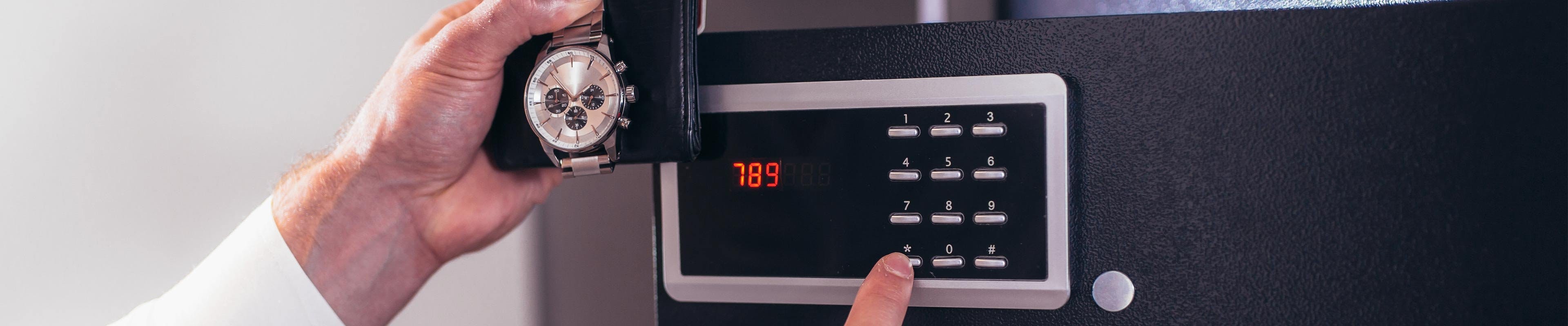 A white man enters a code into a hotel room safe's security panel while holding his expensive watch and wallet.