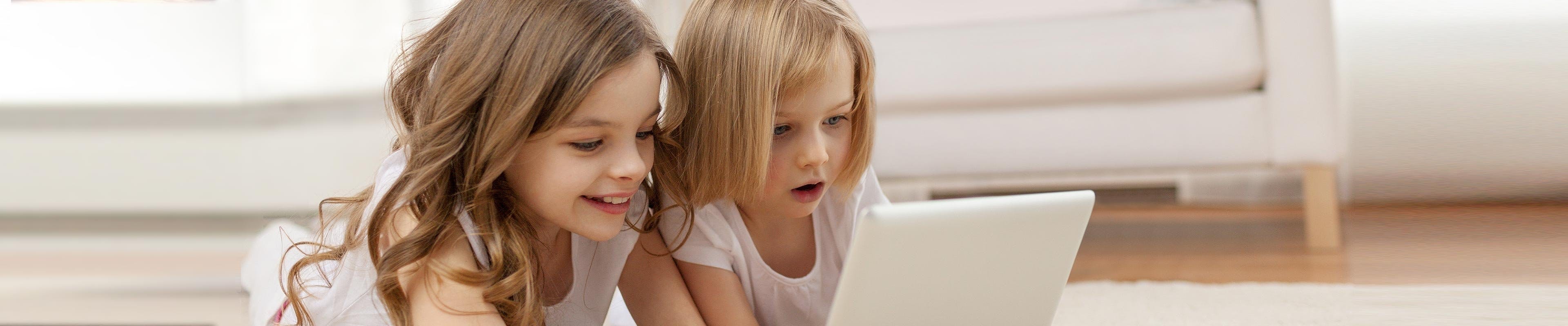 Image of two sisters looking at a tablet while playing in the living room.
