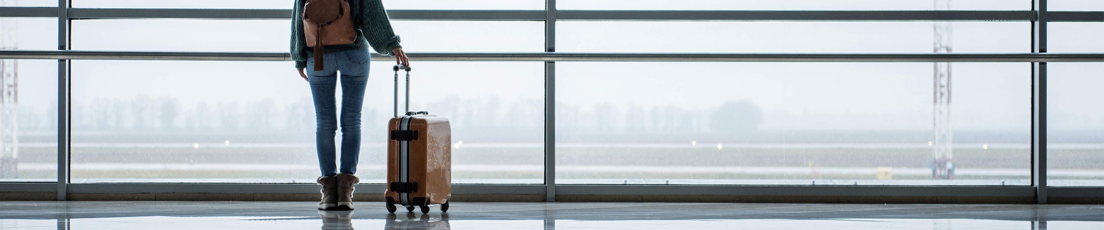 Young woman standing with her back to the camera with a backpack and rolling suitcase looking out at a runway at the airport through a floor-to-ceiling window.