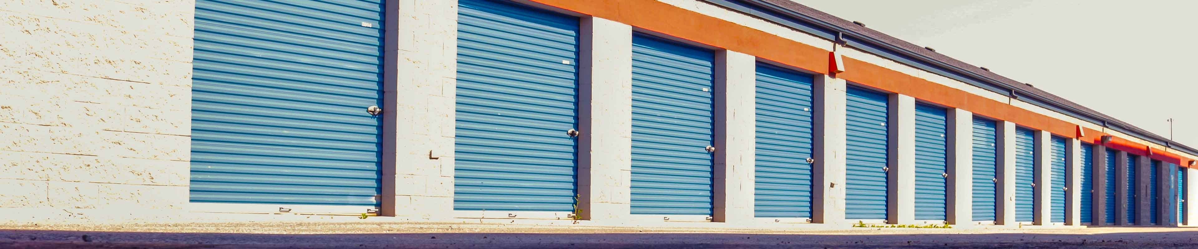 A row of fourteen storage units with sky blue doors and an orange overhang set into a white concrete block building.