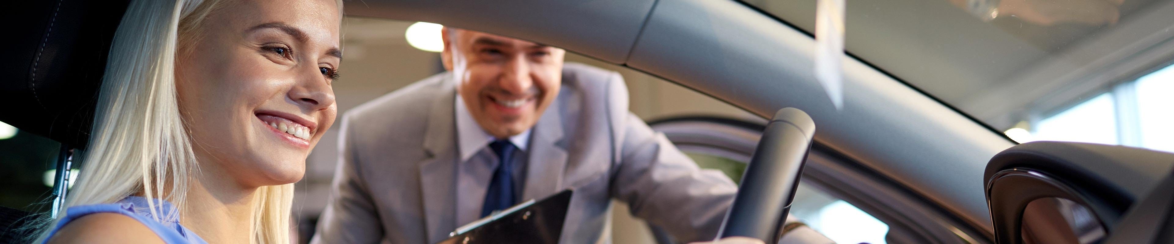 A white blonde woman smiles at her new car while the male salesman stands outside with a clipboard to help her insure the financed car.