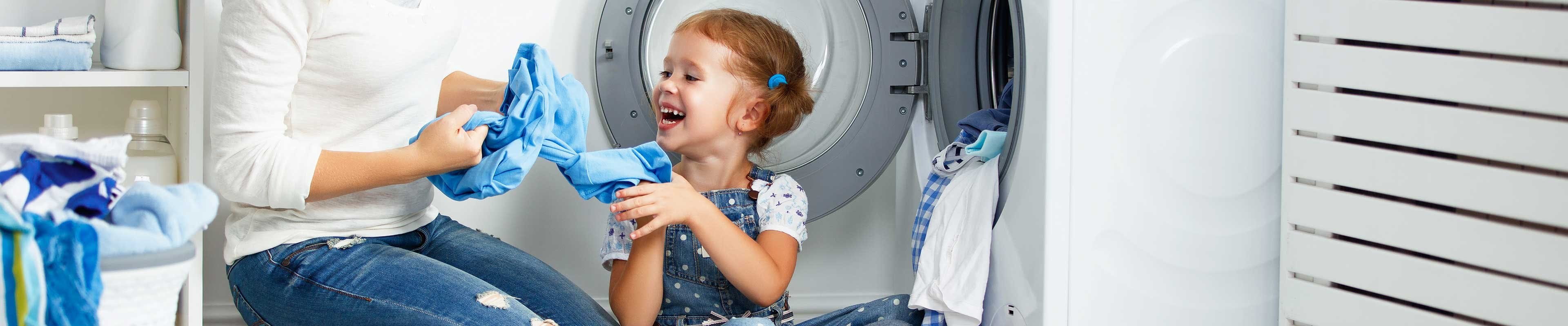 A white mother and her white, red-headed toddler daughter load laundry into a washing machine appliance.