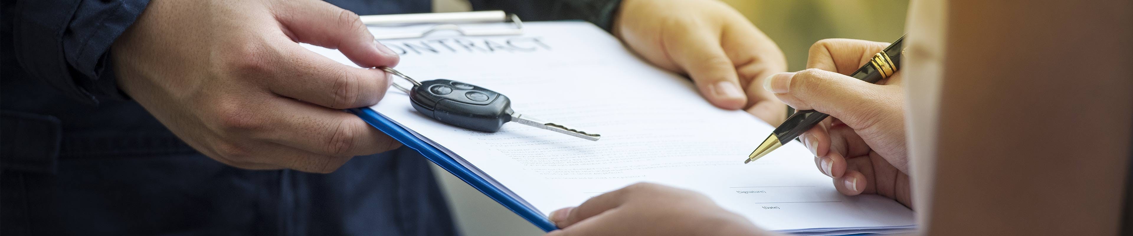 A white woman signs a car lease agreement held on a clipboard by a white man holding her new vehicle's key.