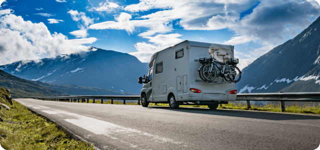 A white rv with bicycles on the back driving on a road with mountains in the background